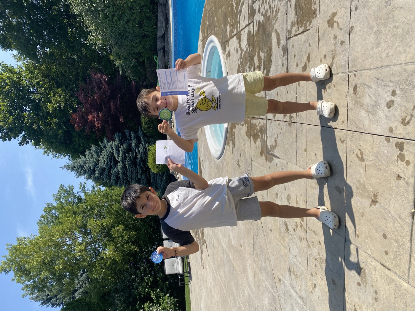 Two young swimmers proudly holding their swim badges and report cards beside the pool