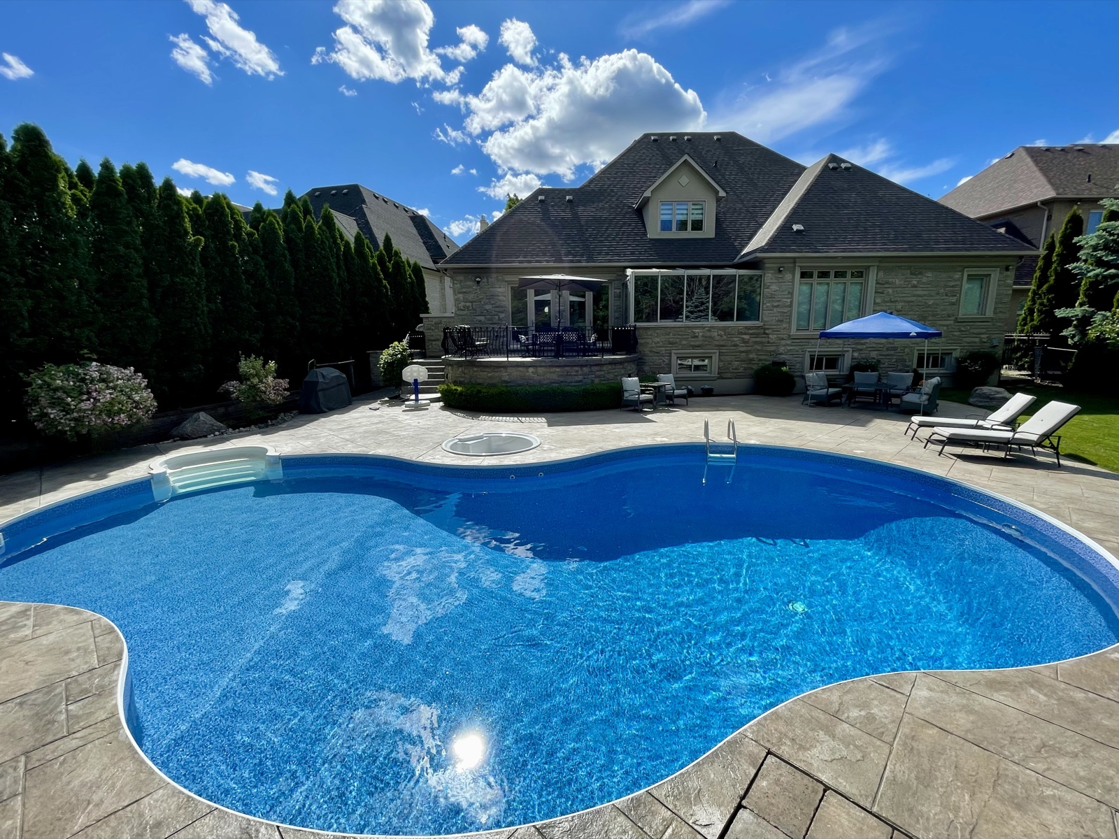 A sunny backyard swimming pool surrounded by a stone patio and lounge chairs