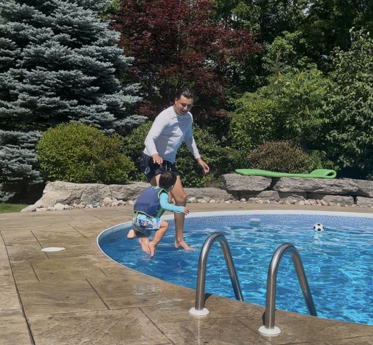 An Ilios instructor and a young swimmer in a life jacket jumping into a backyard pool together